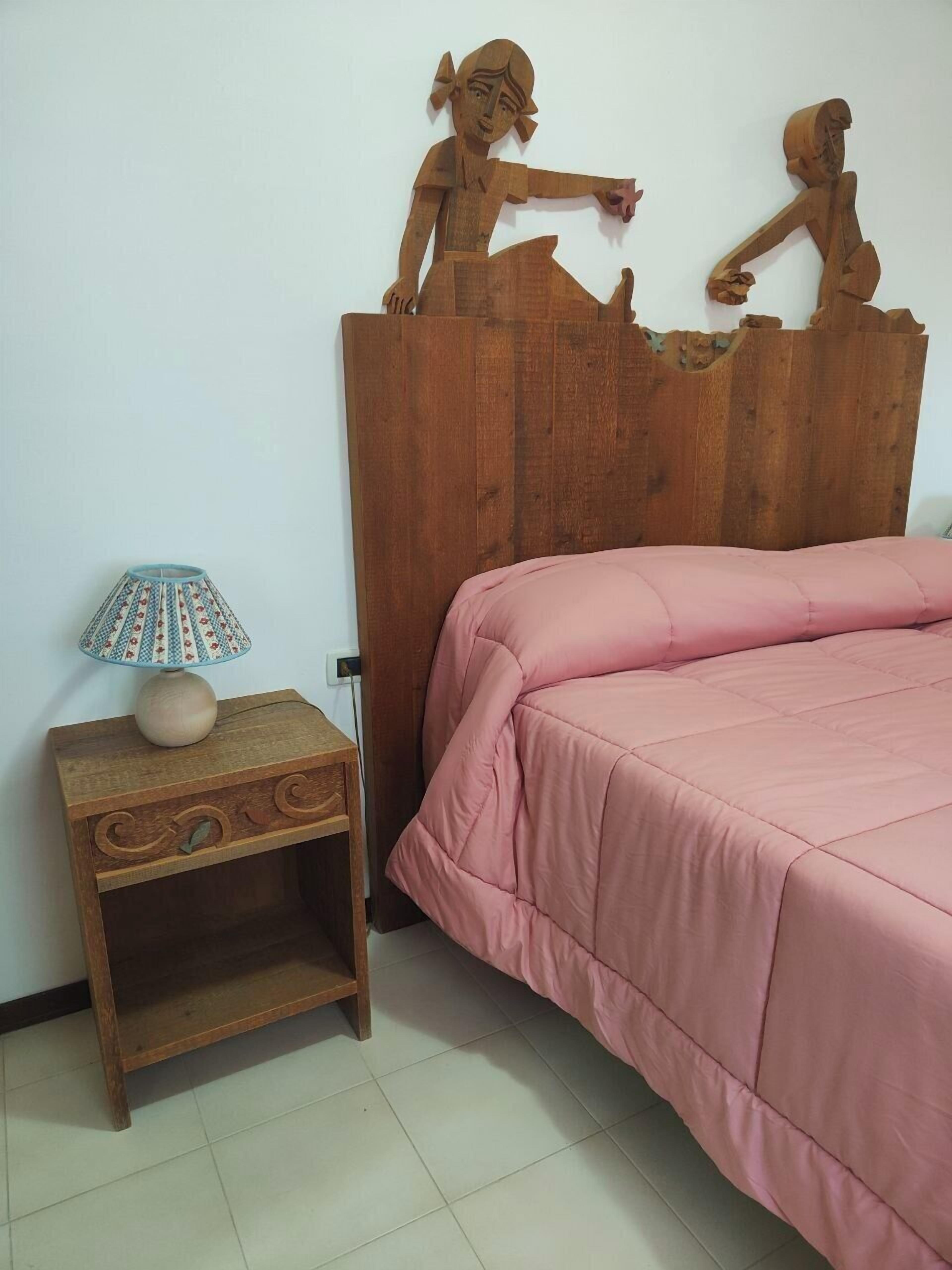 Ein Schlafzimmer mit einem hölzernen Kopfteil, einer rosa Bettdecke, einem Nachttisch mit Lampe und einer kleinen Ablagefläche.