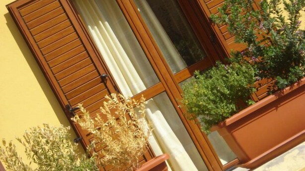 A building with a yellow facade, wooden shutters, and a window with white curtains. There are potted plants on the ground. - Hotel Patti (Golfo Aranci)