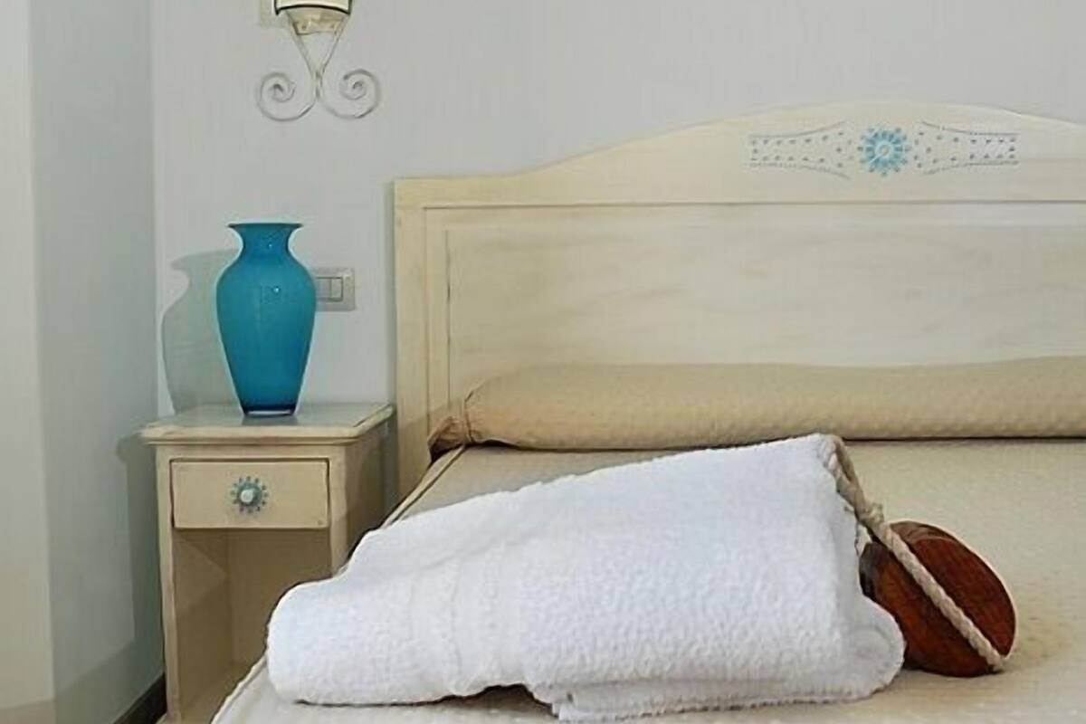 A neatly made bed with a white blanket and a pillow, a small bedside table with a blue vase, and a wall-mounted light fixture.