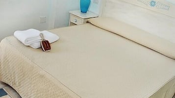 Un lit bien fait, avec une couverture beige et des oreillers, une serviette blanche et un petit vase bleu posé sur une table de chevet.