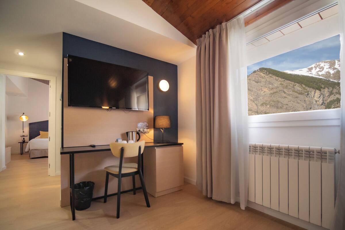 A hotel room with a bed, a desk with a chair, a TV, a window with curtains, and a view of a mountain.