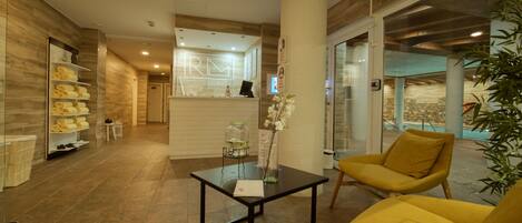 A modern spa lobby with a reception desk, a glass wall, a reception area, a yellow chair, and a glass table with a vase of flowers.
