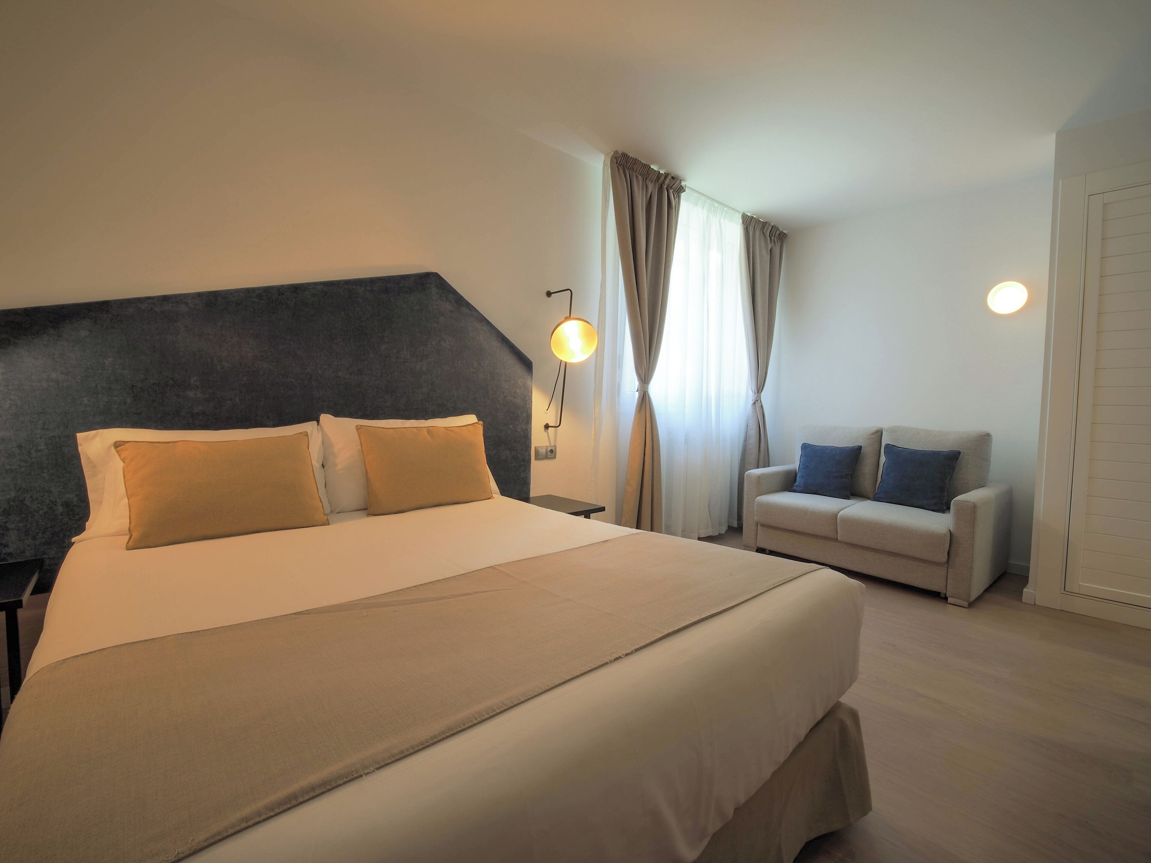 A hotel room with a large bed, a sofa, a wall-mounted lamp, and a window with curtains.
