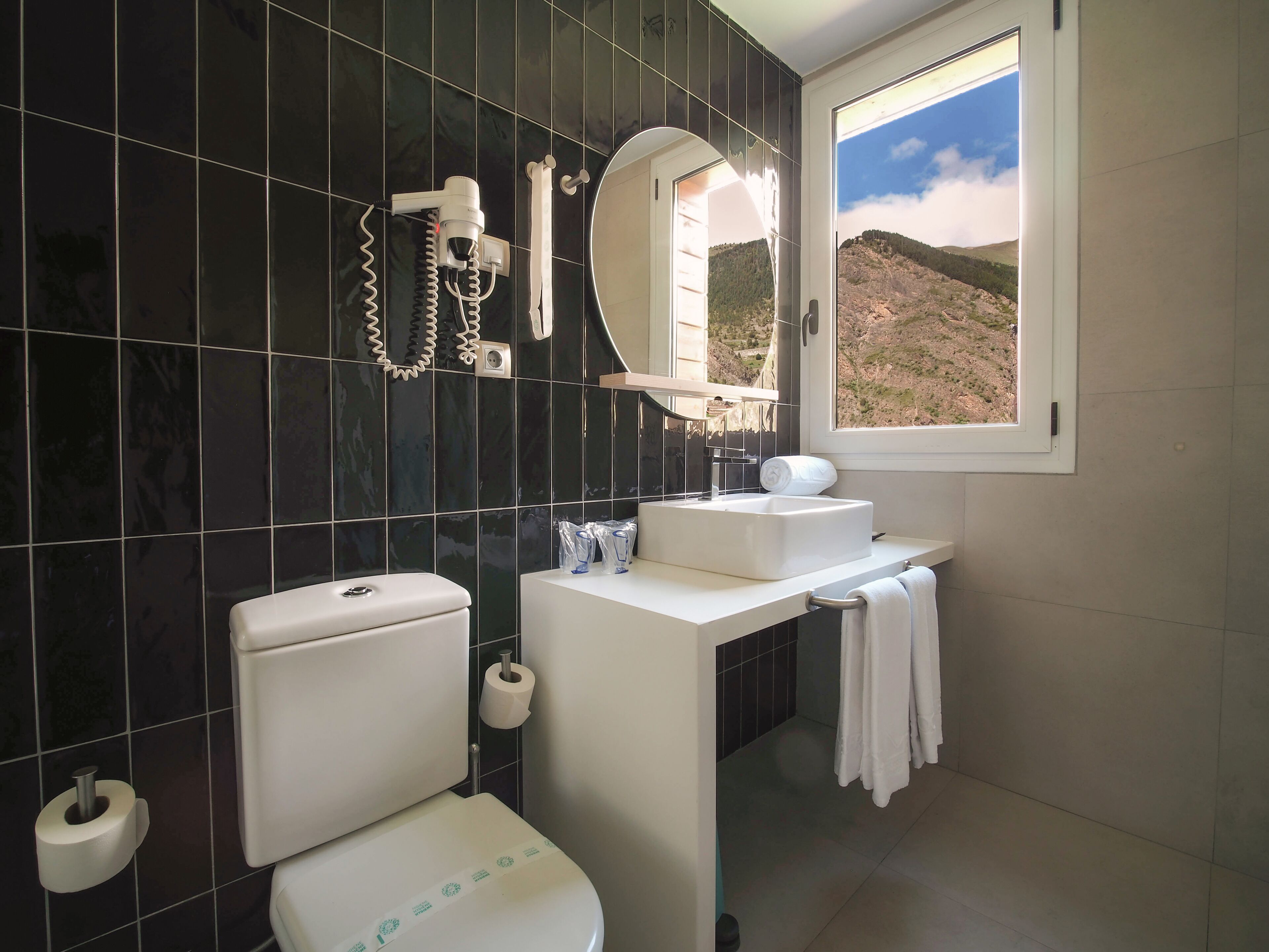 Een badkamer met toilet, wastafel, spiegel en raam met uitzicht op een bergachtig landschap.