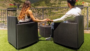 Dos personas sentadas en sillas de mimbre, mirándose, con vistas a un paisaje montañoso y cielo despejado.