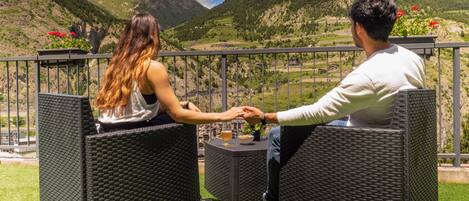 Two people seated on wicker chairs facing each other, overlooking a mountainous landscape with a clear sky.