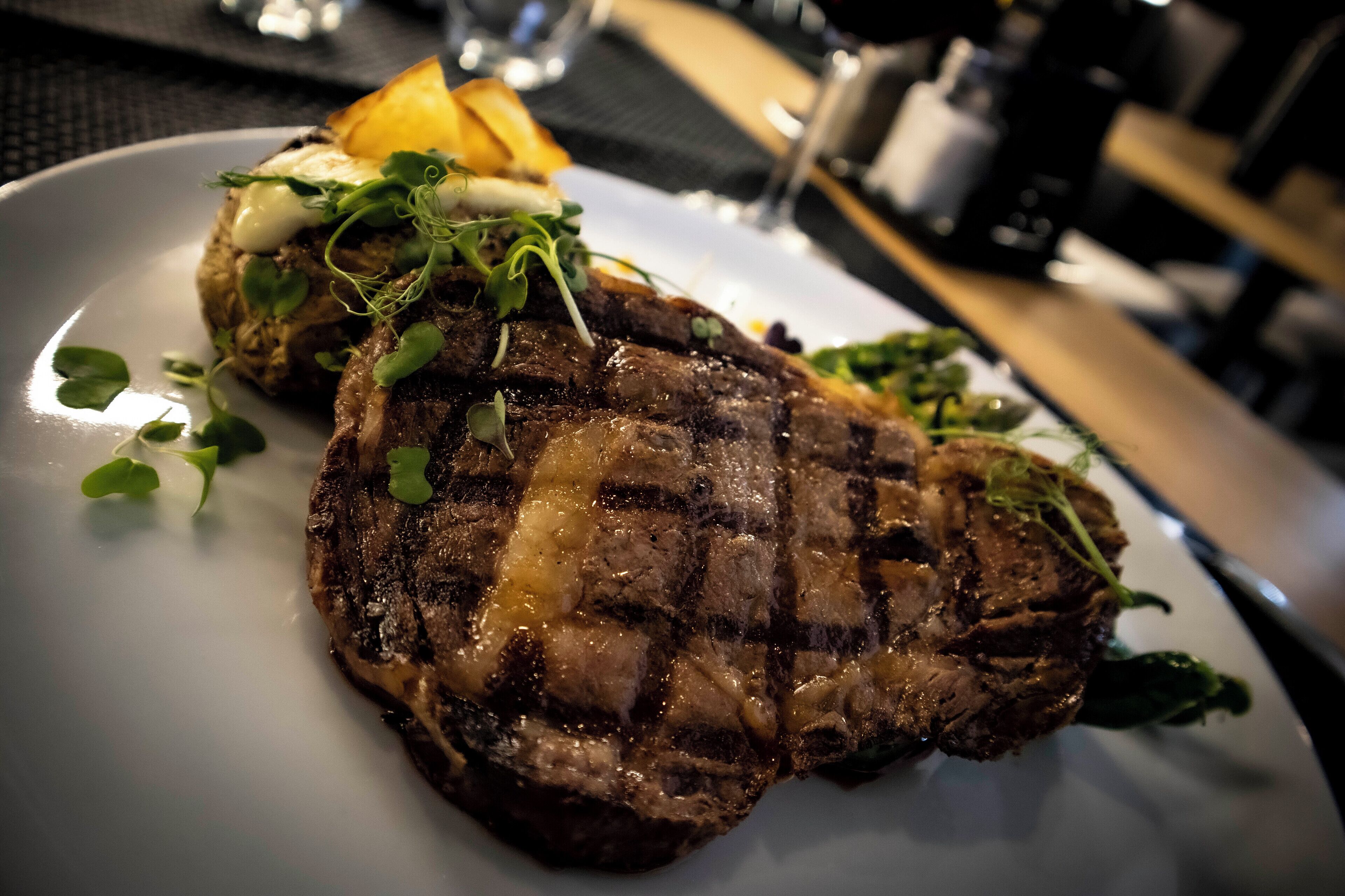 Un bistec con corteza carbonizada, adornado con vegetales verdes y una guarnición de papas asadas.