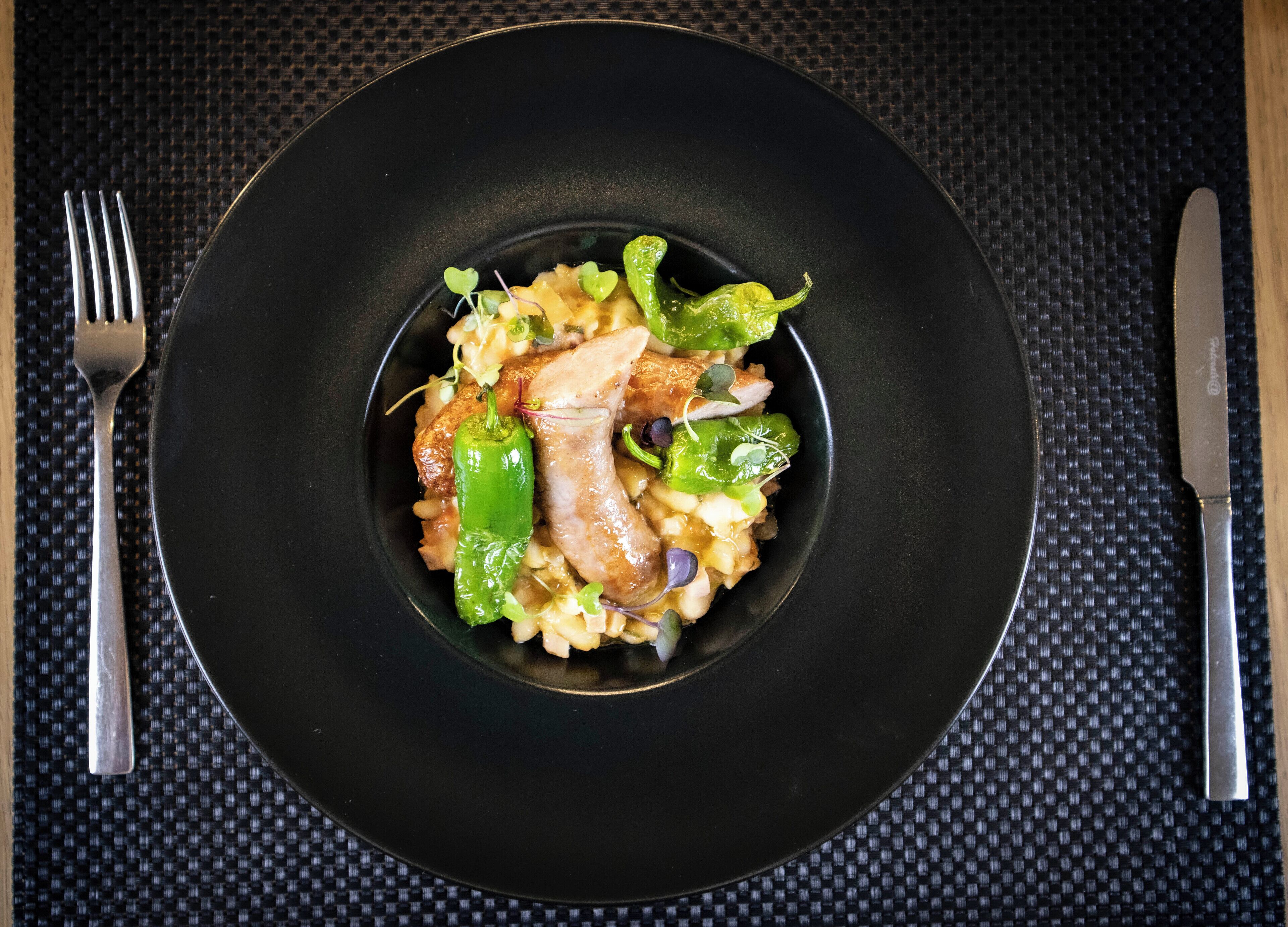 A black plate with a meal consisting of pasta, green peppers, and a piece of meat, placed on a textured tablecloth.