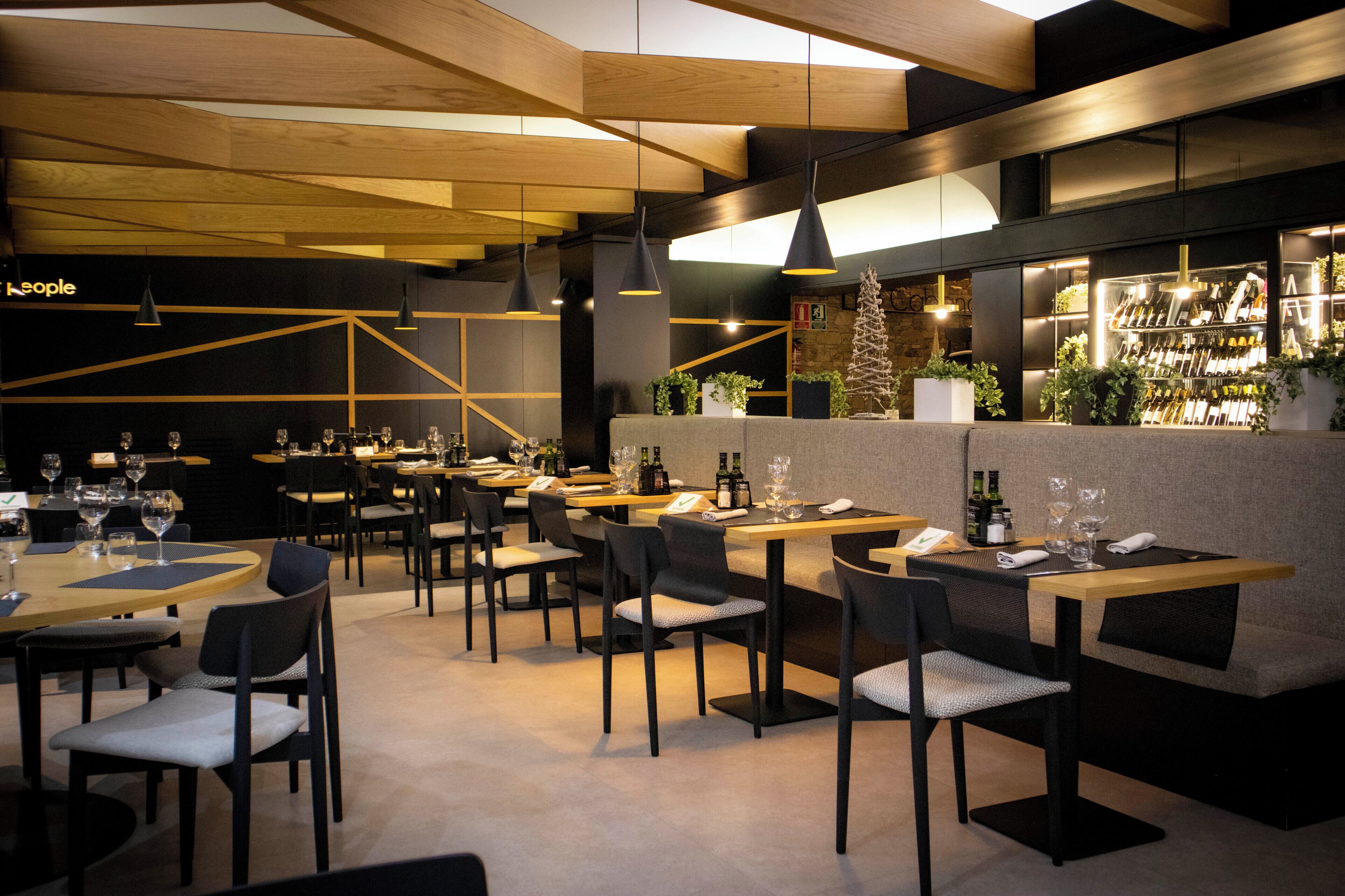 Un restaurant modern amb bigues de sostre de fusta, llums penjants i una zona de bar amb ampolles de vi i vasos.