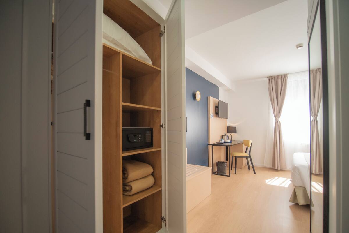 A modern room with a wooden wardrobe, a desk with a chair, a small table with a lamp, and a bed with curtains.