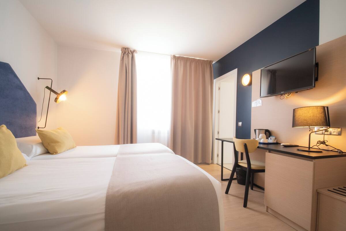 A hotel room with two beds, a desk, a chair, a TV, and a lamp.
