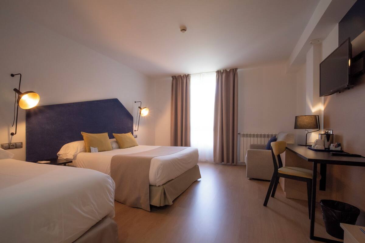 A hotel room with two beds, a desk, a chair, a TV, and a window with curtains.