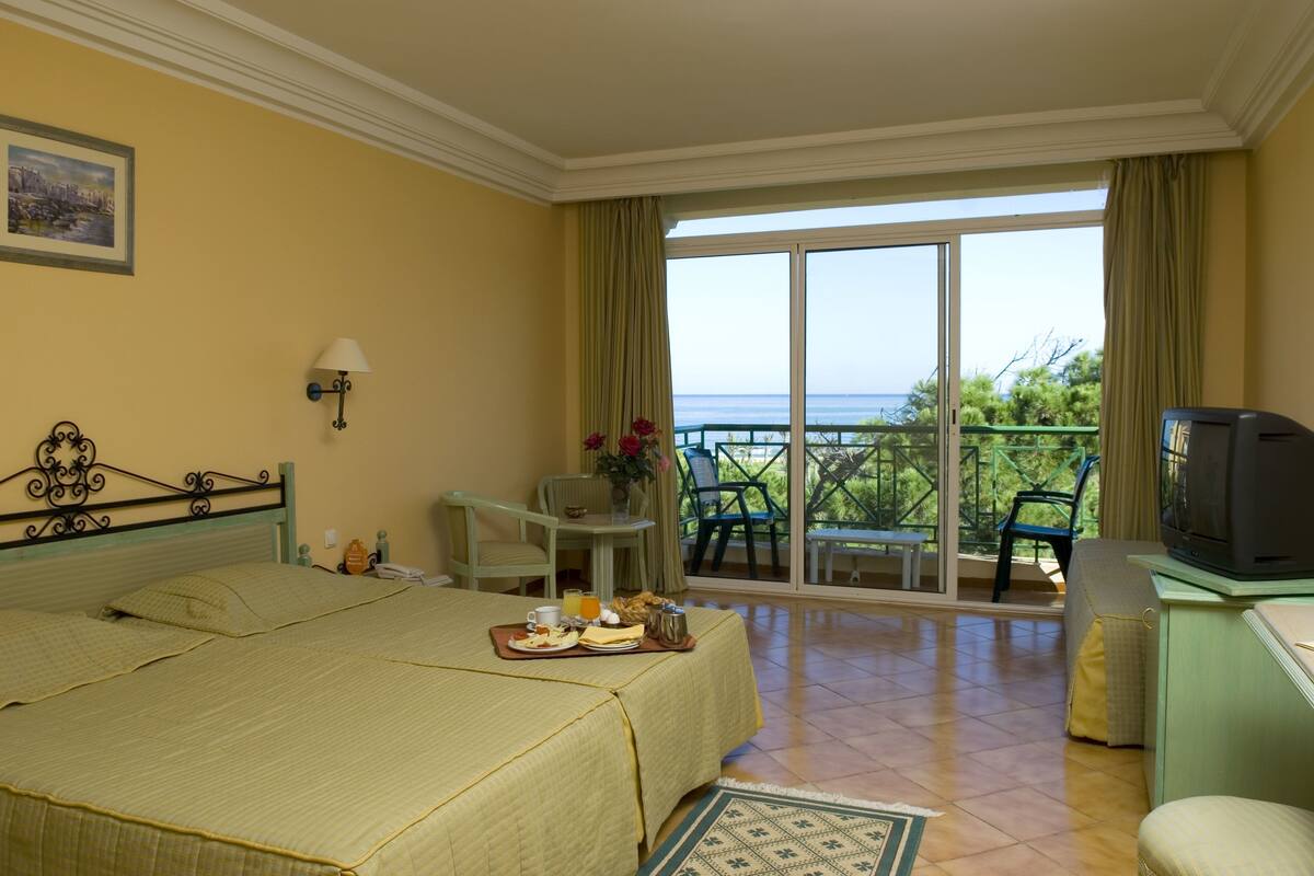 A hotel room with a bed, a desk with a chair, a television, and a balcony with a view of the sea.
