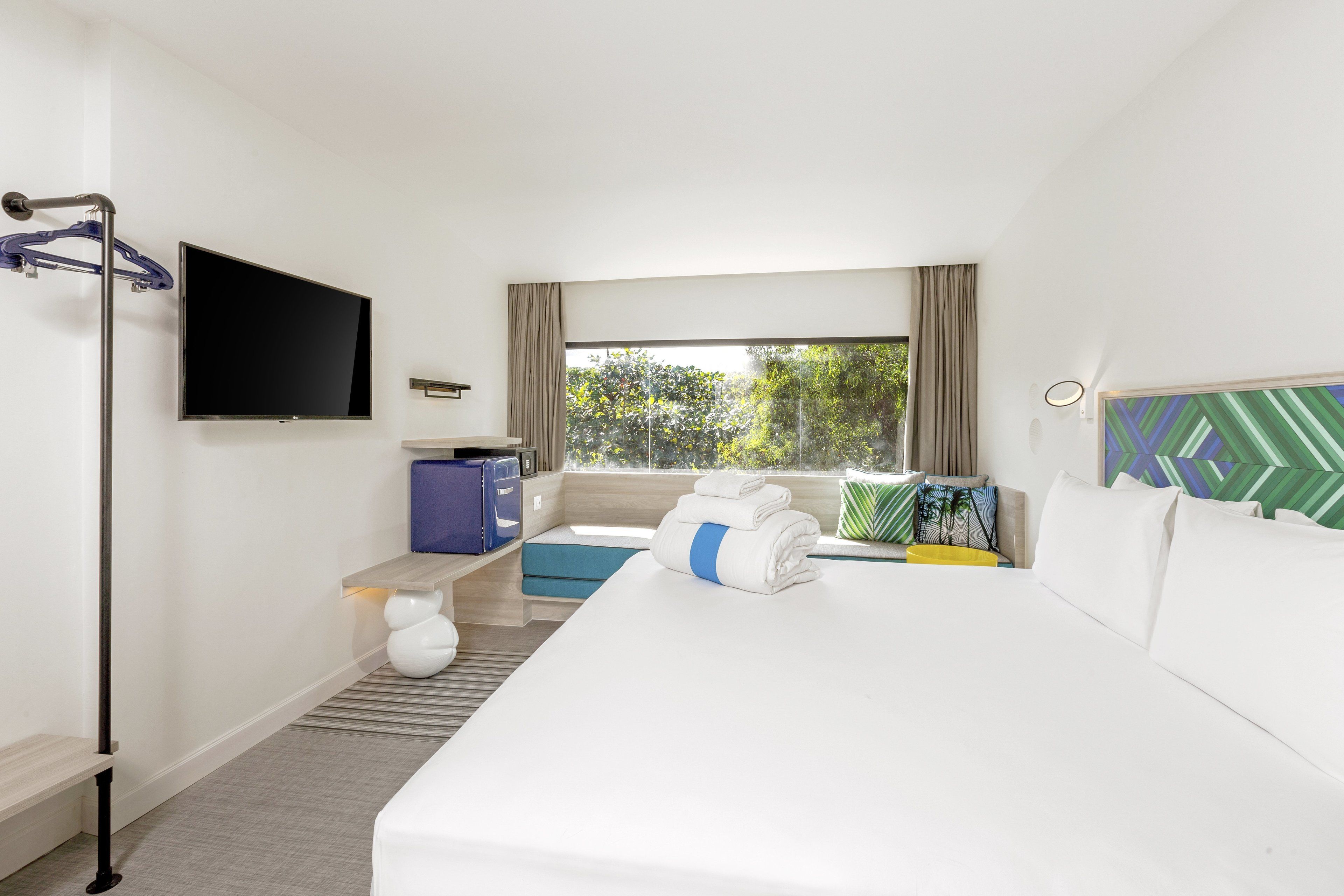 A hotel room with a large bed, a flat-screen TV, a desk with a chair, a window with curtains, and a view of greenery outside.