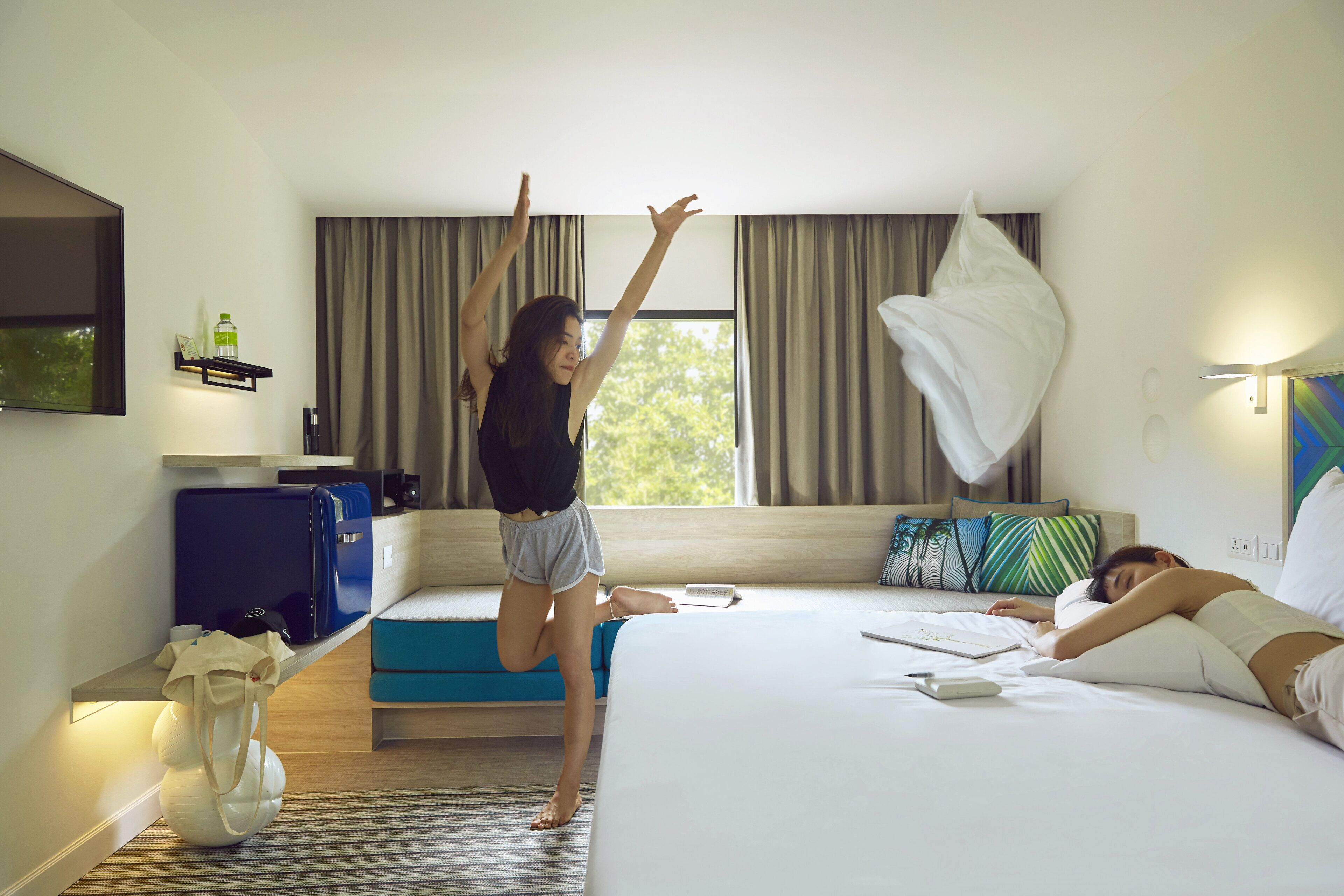 A hotel room with a bed, a blue suitcase, a white vase, and a window with curtains.