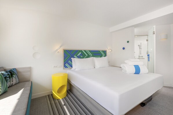 Una habitación de hotel moderna con una cama grande, un cabecero con diseño geométrico, un taburete pequeño amarillo y una alfombra a rayas.