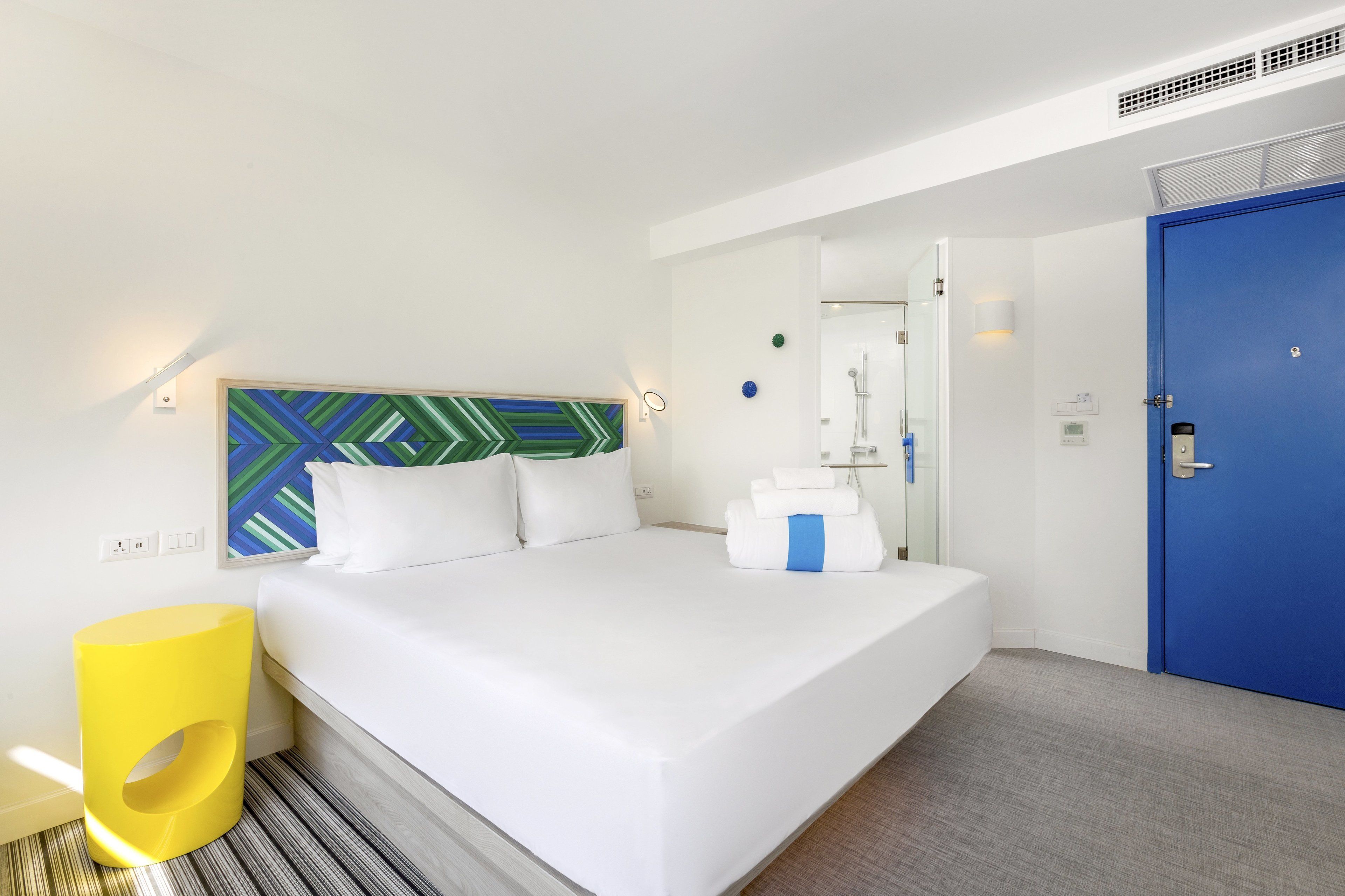 A hotel room with a bed, a blue door, a mirror, and a wall-mounted light fixture.