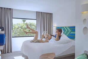 A modern hotel room with a large bed, a desk with a suitcase, and a view of trees through the window. - COSI Samui Chaweng Beach (Koh Samui)