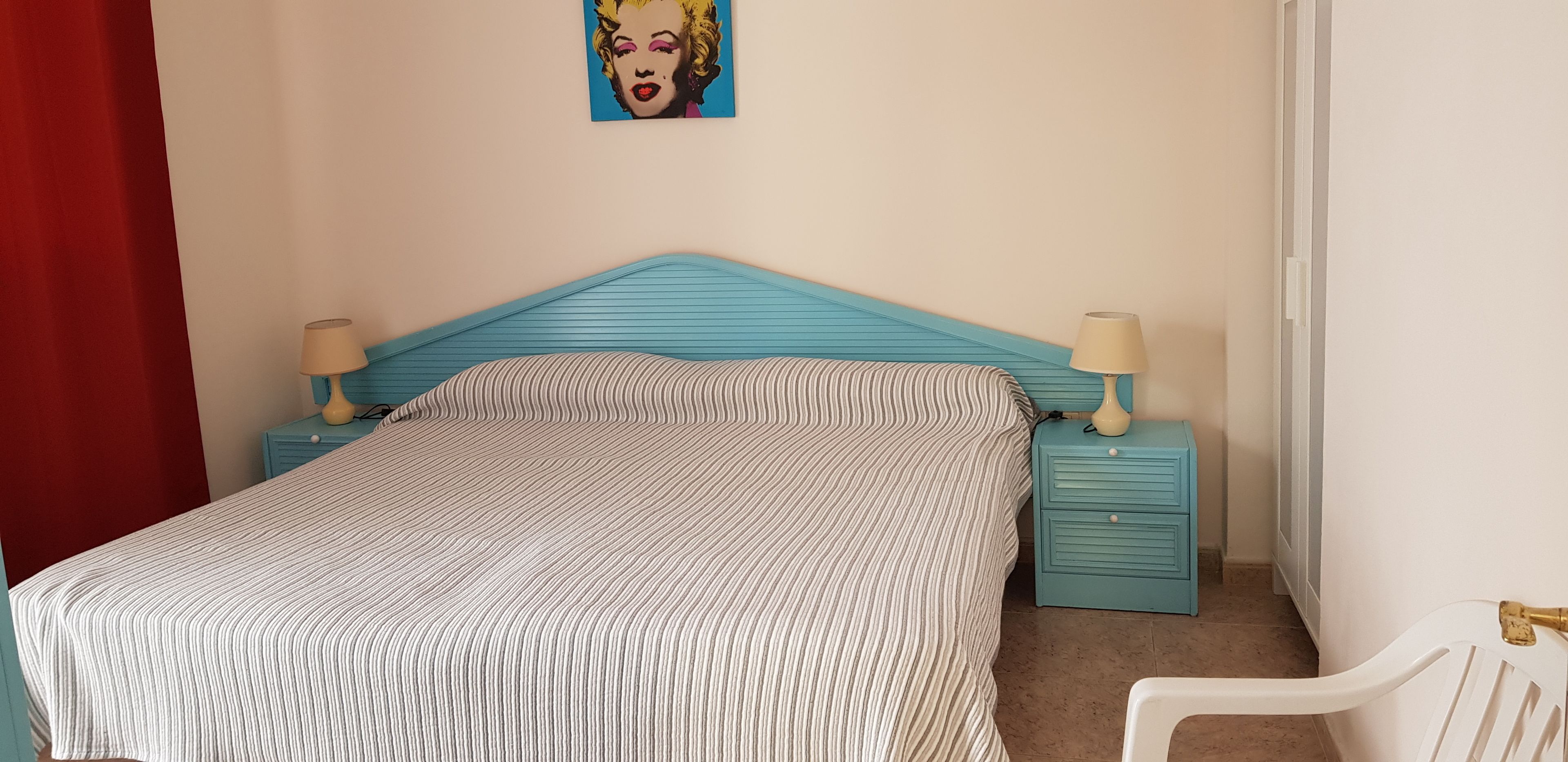 A bedroom with a striped bedspread, a blue headboard, two bedside tables with lamps, a red curtain, and a wall painting.