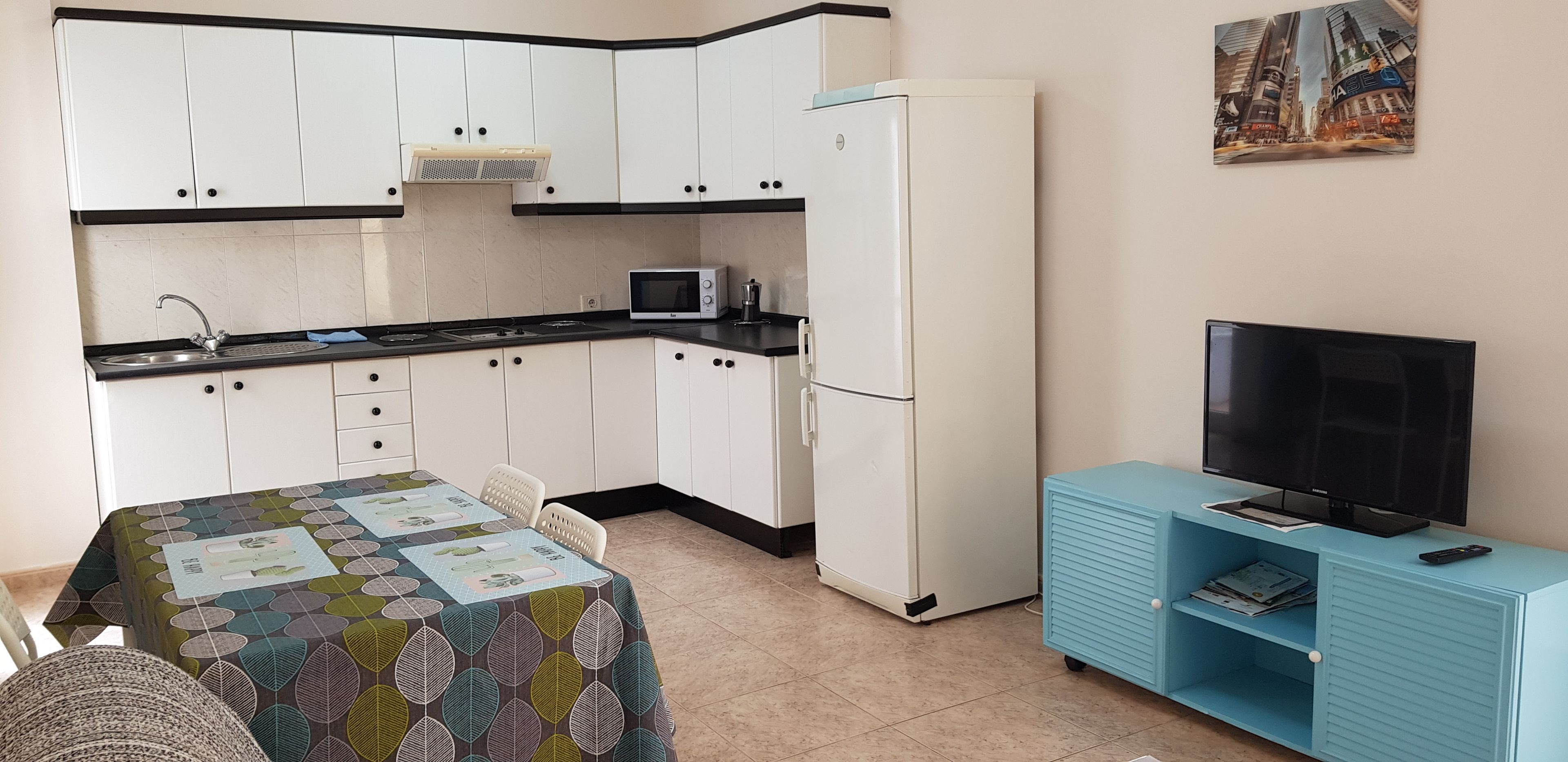 Een kleine keuken met witte kastjes, een magnetron, een koelkast en een blauwe entertainmentkast met een televisie.