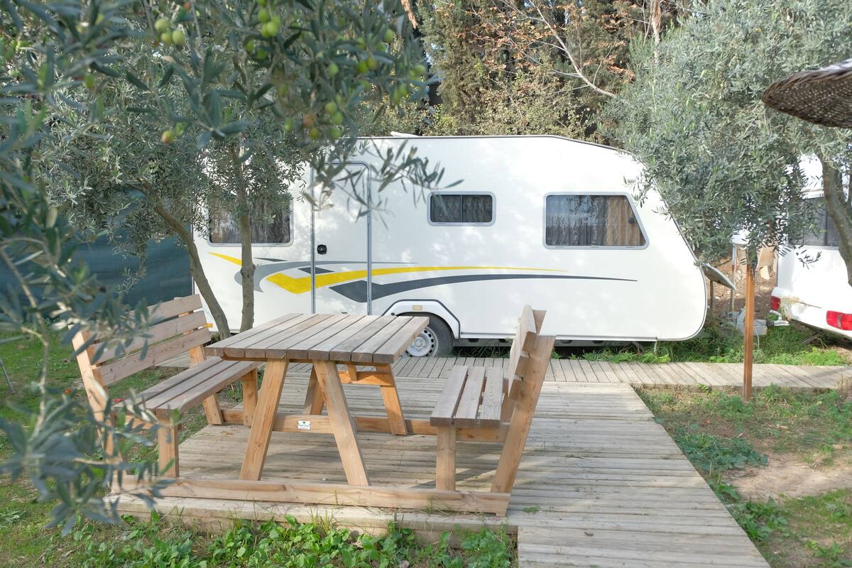 Una caravana blanca estacionada en un área boscosa con una mesa de picnic de madera y bancos.