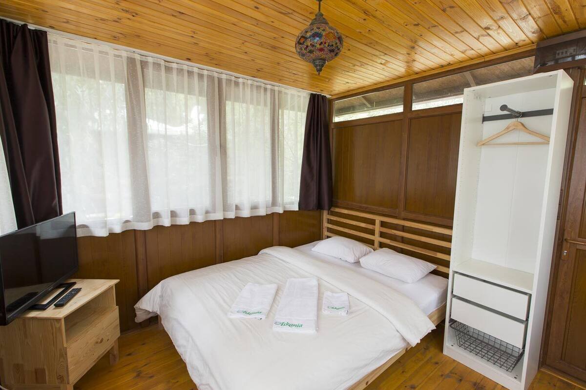 Un dormitorio con techo de madera, una cama con ropa blanca, un televisor sobre un mueble de madera y un armario con barra para colgar.