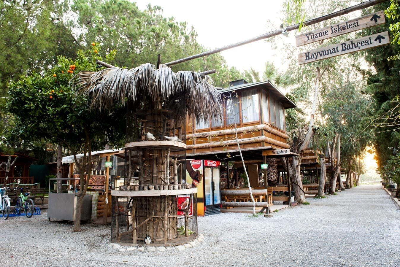 A rustic wooden building with a thatched roof, a Coca-Cola sign, and a signpost indicating directions to 'Yuzme İskeleşi' and 'Hayvanat Bahçesi'.
