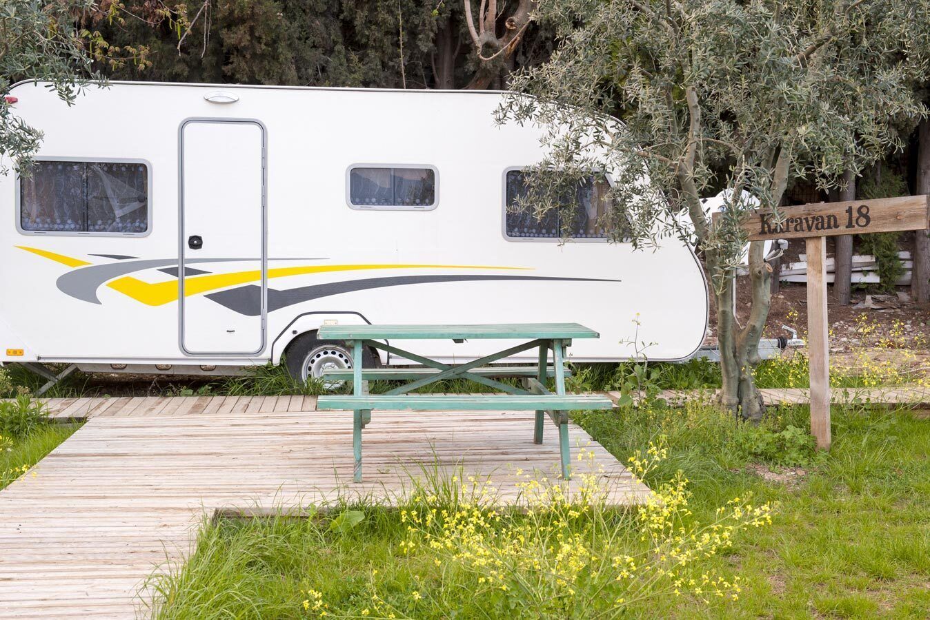 Una caravana con una mesa de picnic al frente, rodeada de vegetación y un letrero de madera que dice 'Koravan 18'.