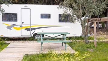 Una caravana con una mesa de picnic al frente, rodeada de vegetación y un letrero de madera que dice 'Koravan 18'.