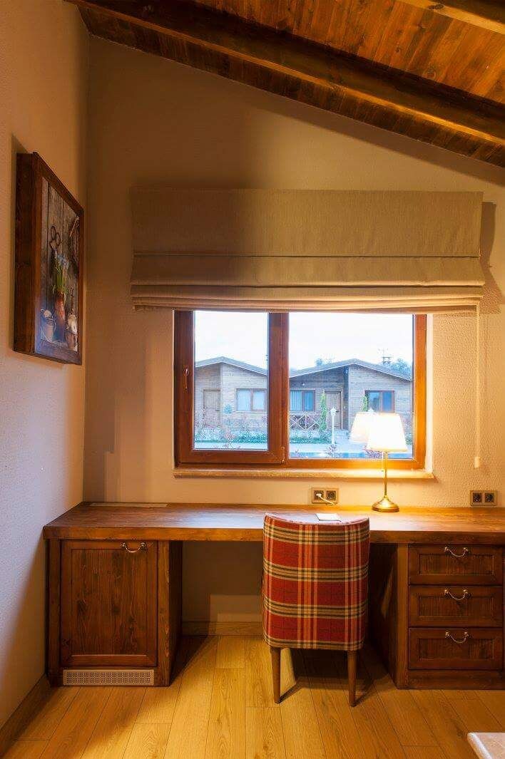 A wooden desk with a plaid chair, a window with a view of buildings, and a lamp on the desk.