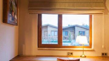 A wooden desk with a plaid chair, a window with a view of buildings, and a lamp on the desk.