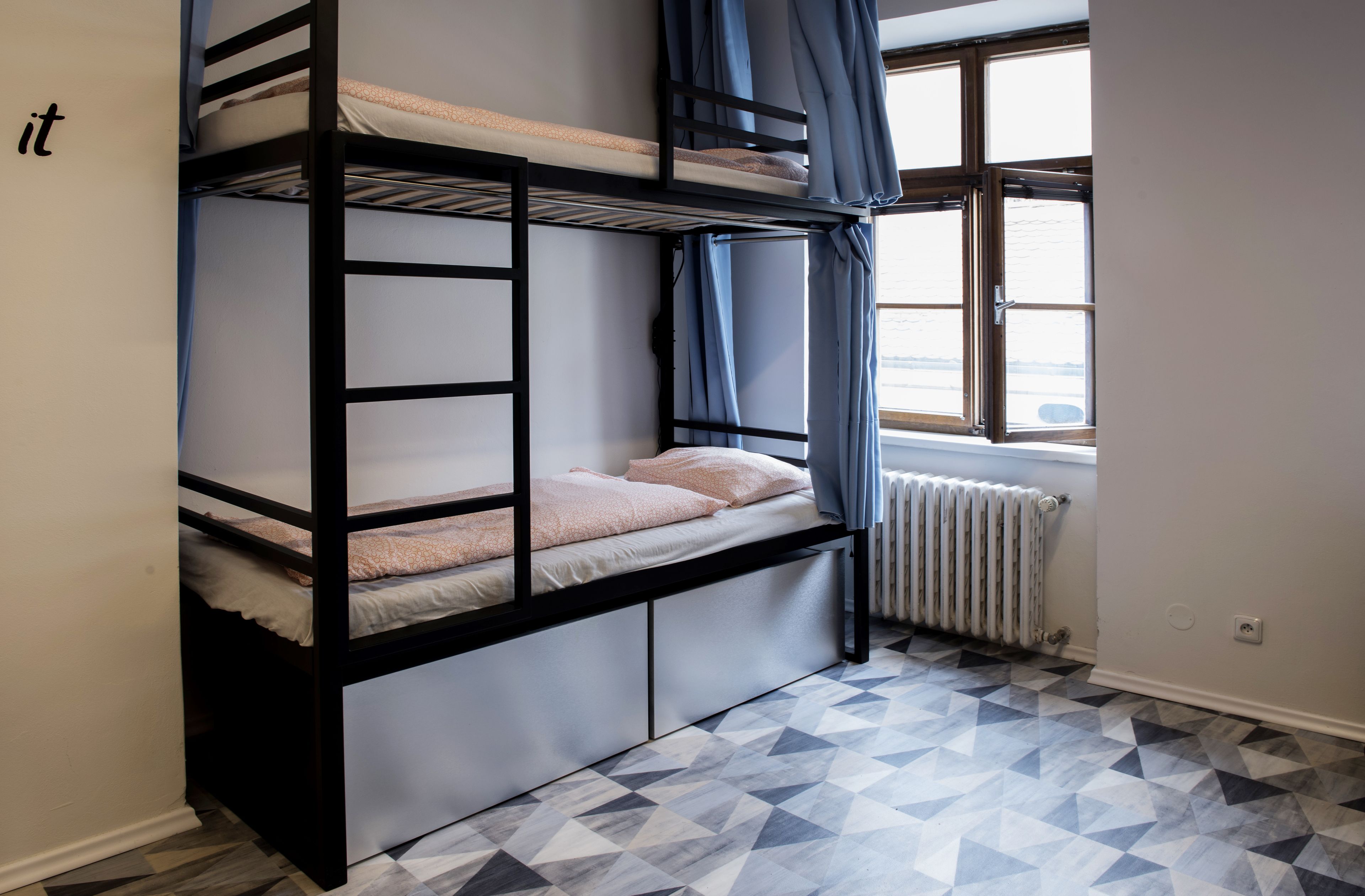 Habitación con literas, suelo de diseño geométrico, ventana con cortinas y un radiador.