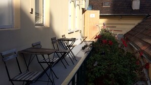 Un balcon avec une table et des chaises, un pot de fleurs avec des fleurs rouges, et un bâtiment avec un toit rouge en arrière-plan.