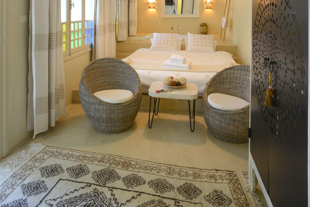 A bedroom with a bed, two wicker chairs, a nightstand, a mirror, and a decorative wall panel.