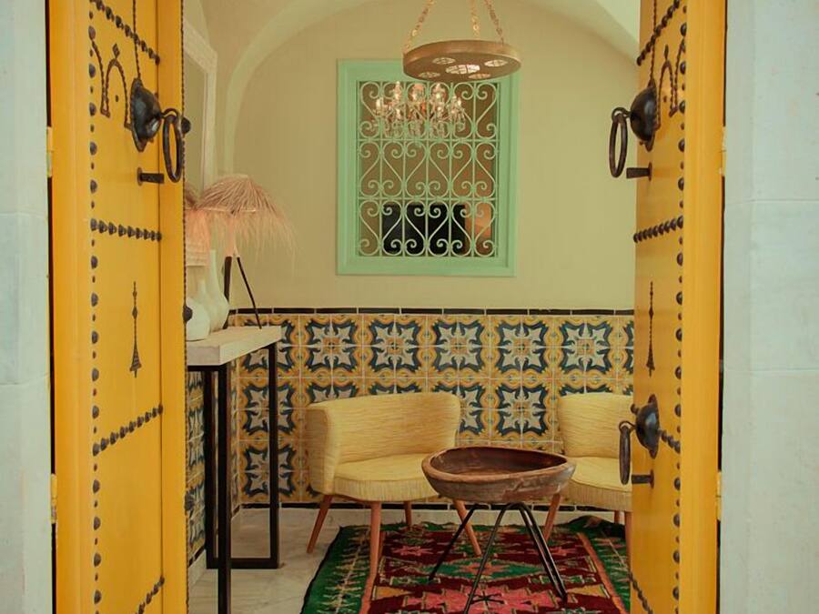 A room with a yellow door, a green window with decorative ironwork, a patterned wall, a sofa, a small table, and a hanging chandelier.