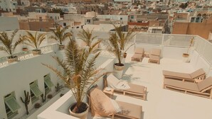 En takterrass med solstolar och krukväxter, med utsikt över ett tättbebyggt stadskvarter med traditionell arkitektur.