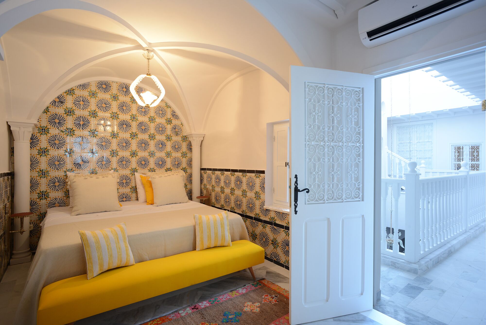 Un dormitorio con cama, un banco amarillo, papel tapiz estampado, una araña y un balcón con barandilla.