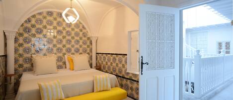 Une chambre à coucher avec un lit, un banc jaune, un papier peint à motifs, un lustre et un balcon doté d’une balustrade.