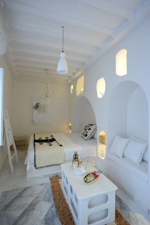 A minimalist white room with a bed, a small table, and a sofa. There are wall sconces providing light.