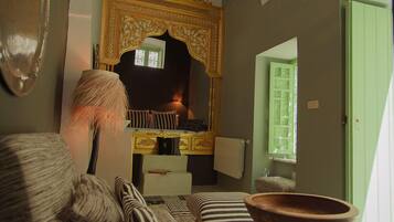 Una habitación con una cama, una silla de madera, una mesita y un espejo con un marco decorativo.