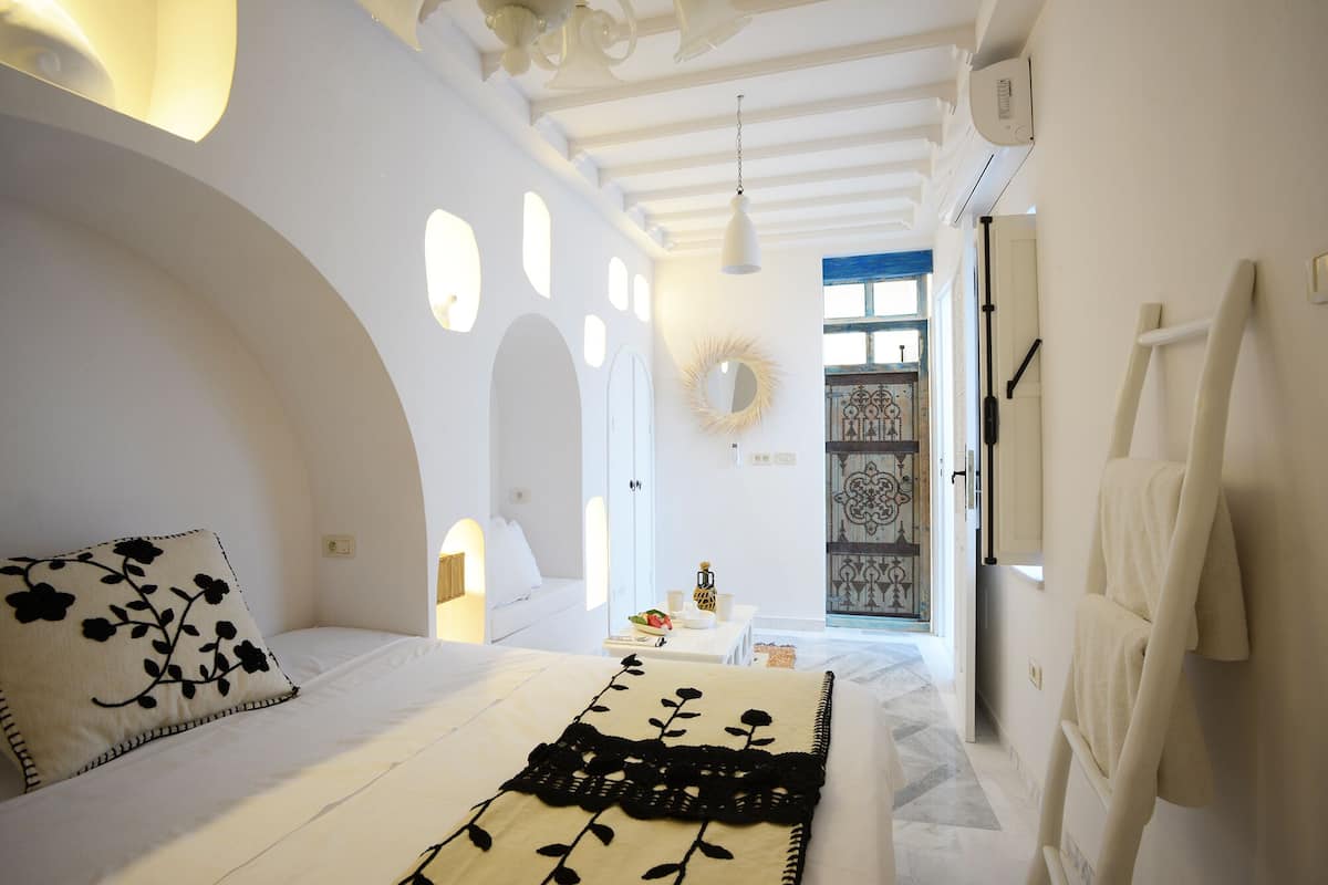 A bedroom with a bed, a bedside table, a mirror, and a view of the outside.