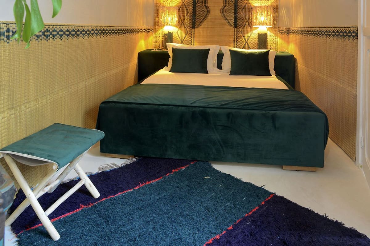 A neatly arranged bedroom with a bed, bedside lamps, a hanging light fixture, a small table, and a large plant.