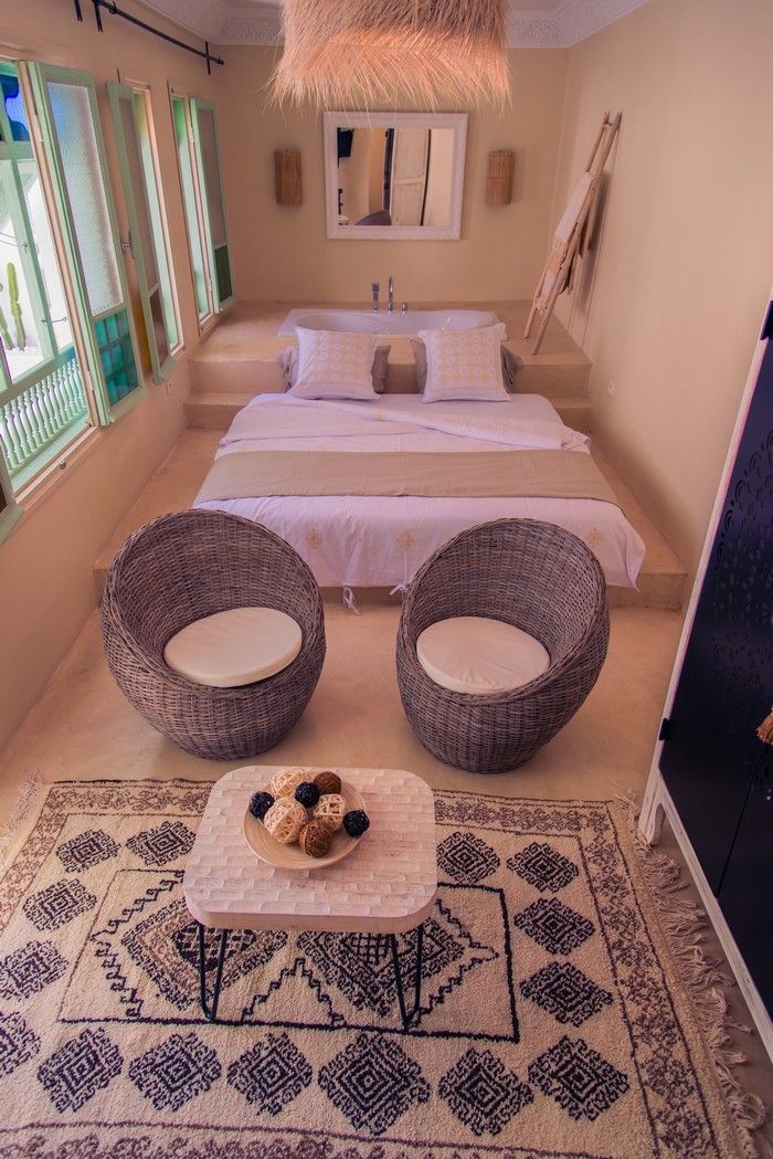 A bedroom with a bed, two wicker chairs, a small table with decorative items, and a patterned rug.