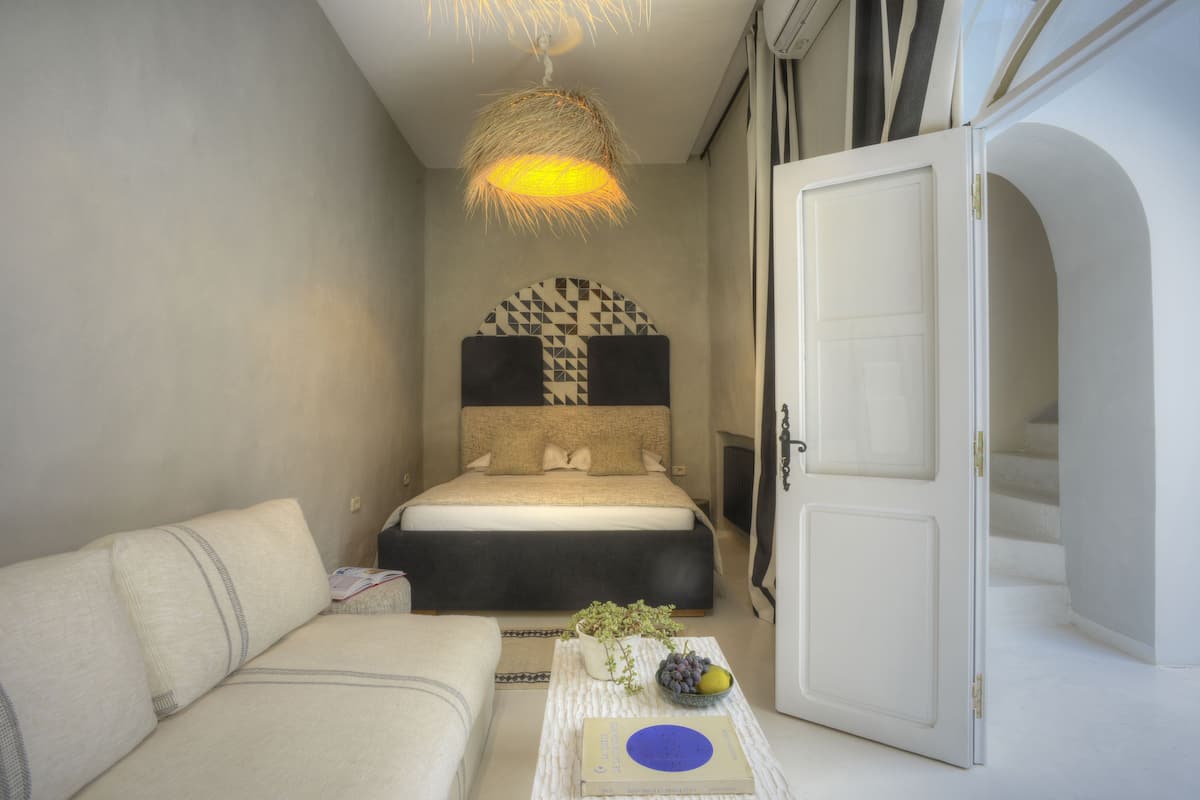 A bedroom with a bed, a sofa, a bedside table with a book and a bowl of fruit, and a hanging light fixture.