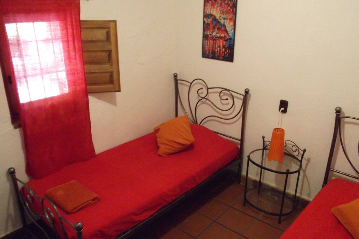 Una habitación pequeña con una cama individual, colcha roja, puerta de madera, cortina roja, una mesita con lámpara y un cuadro en la pared.