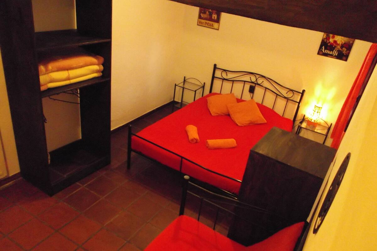 Una habitación pequeña con cama individual, colcha roja, cabecero de metal y repisas con toallas dobladas.