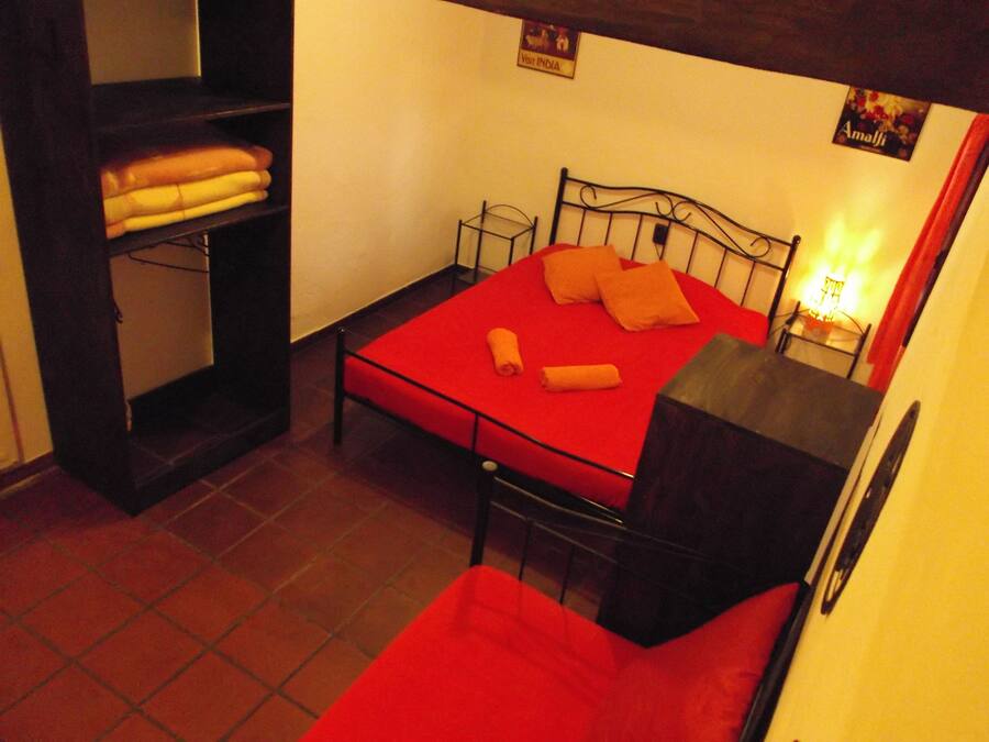 Una habitación pequeña con cama individual, colcha roja, cabecero de metal y repisas con toallas dobladas.