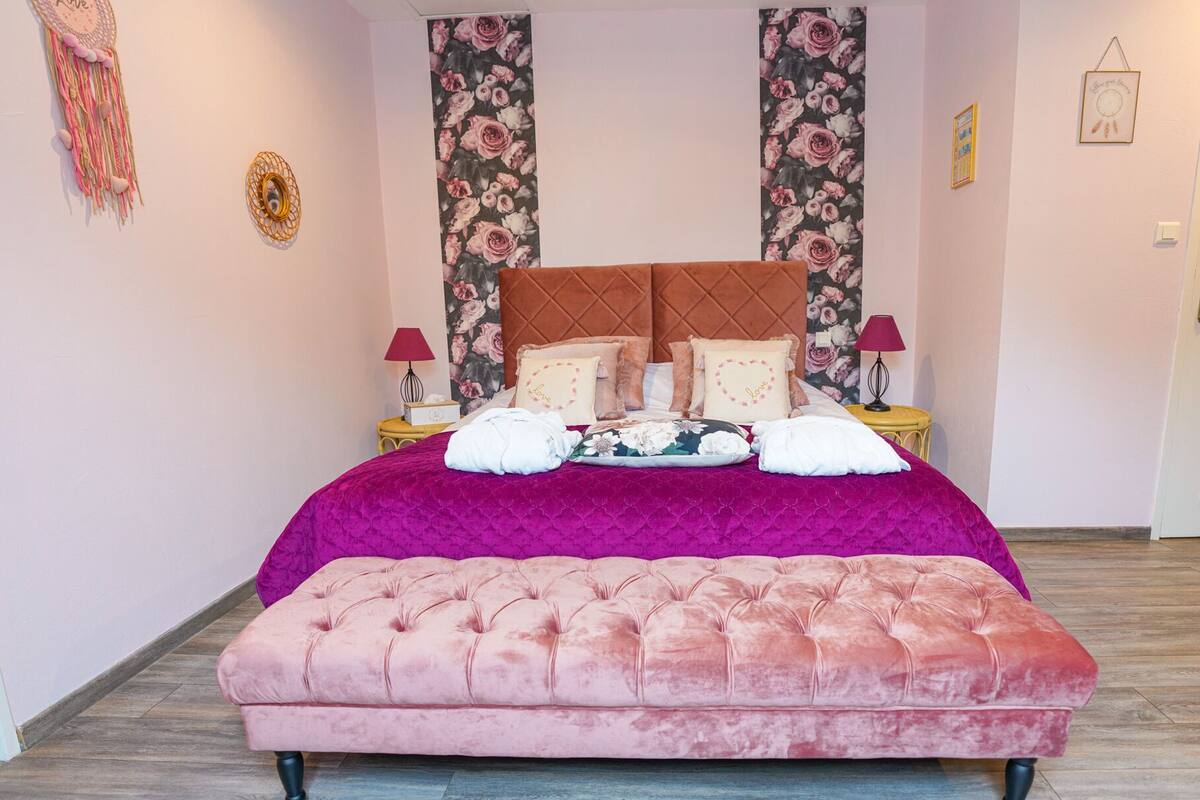 Une chambre avec un grand lit, un banc recouvert d’un tissu rose, un papier peint à motifs floraux et deux tables de chevet avec des lampes.