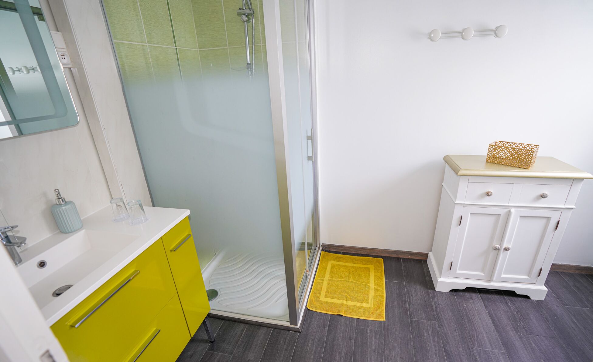 Un baño con cabina de ducha de vidrio, un mueble amarillo, un mueble blanco con una caja decorativa y un felpudo amarillo.