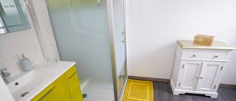 Un baño con cabina de ducha de vidrio, un mueble amarillo, un mueble blanco con una caja decorativa y un felpudo amarillo.